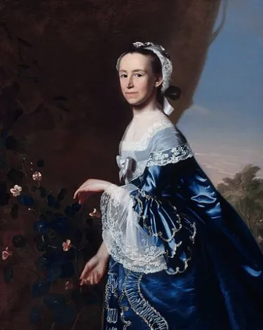 A woman in a blue colonial-era gown looks at the viewer while standing next to flowers. 